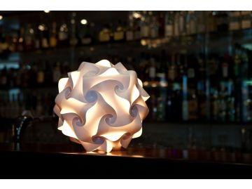 Das Sphere Lichtobjekt (Ø 27 cm) der Lampenmanufaktur Freiburg lässt den Bartresen in geometrischem Weiß erstrahlen, mit sanft verschwommenen Flaschen im Hintergrund.