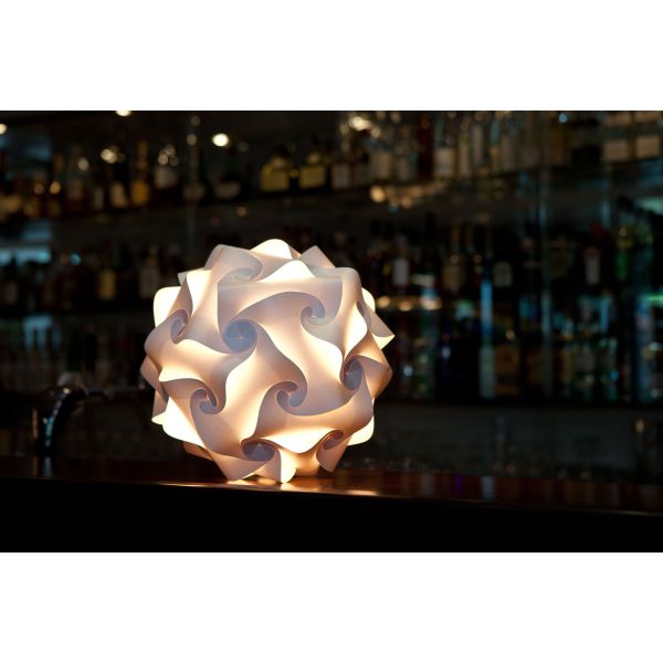 Das Sphere Lichtobjekt (Ø 27 cm) der Lampenmanufaktur Freiburg lässt den Bartresen in geometrischem Weiß erstrahlen, mit sanft verschwommenen Flaschen im Hintergrund.