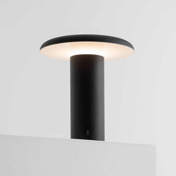 Die Takku Tischleuchte (schwarz) ist eine moderne, minimalistische, kabellose Design-Leuchte mit einer breiten, runden Leuchtfläche, die sanftes weißes Licht ausstrahlt. Sie wird auf einer schlichten weißen Fläche vor einem hellgrauen Hintergrund abgebild
