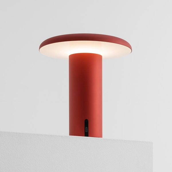 Die Takku Tischleuchte (rot) ist eine moderne, kabellose Designleuchte von Artemide mit einem zylindrischen Sockel und einem breiten Schirm, die auf einer weißen Fläche vor einem schlichten weißen Hintergrund abgebildet ist und ein sanftes Licht ausstrahl