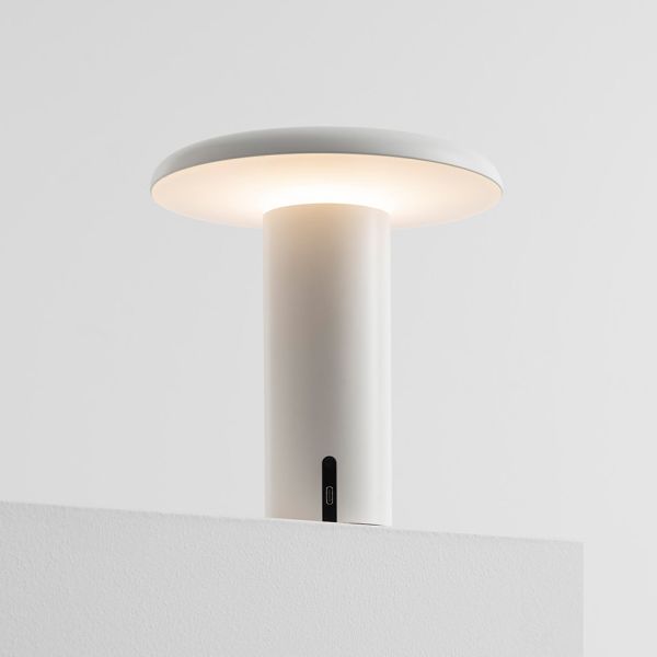 Minimalistische Takku-Tischleuchte (weiß): Kabelloses Design, zylindrischer Sockel, breite Leuchtfläche und sanftes Leuchten – stilvoll in Weiß vor grauem Hintergrund.