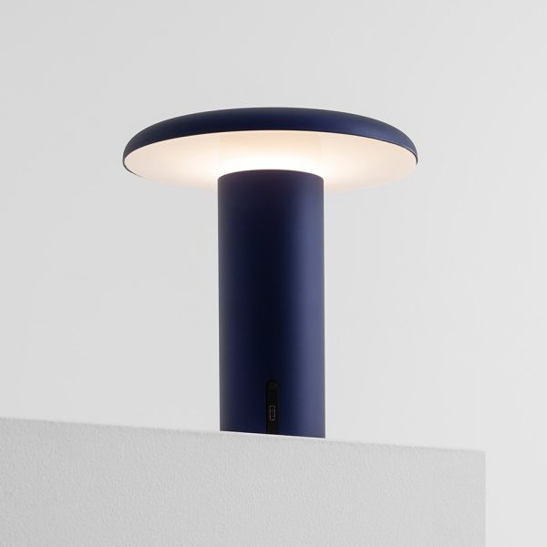 Die Takku Tischleuchte, eine moderne kabellose Lampe mit einem dunkelblauen, zylindrischen Sockel und einem breiten Lampenschirm, strahlt sanftes weißes Licht aus und kommt auf einer weißen Oberfläche vor einem hellgrauen Hintergrund besonders elegant zur