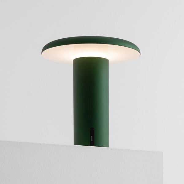 Die grüne Takku-Tischleuchte von Artemide strahlt sanftes weißes Licht aus ihrer flachen Oberseite aus und steht als kabellose Tischleuchte auf einer weißen Fläche vor grauem Hintergrund.
